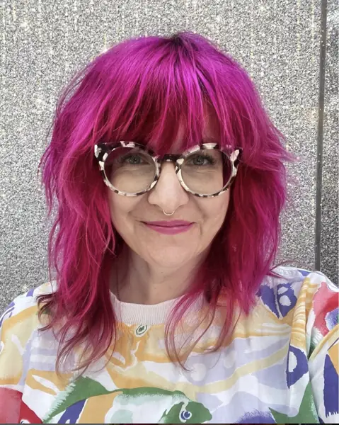 Jen Larsen, smiling, with bright magenta hair and patterned glasses